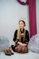 Beautiful young Asian woman in traditional Javanese bridal attire, smiling gracefully with a calm and confident expression, sitting in a studio setting with cultural accessories. photo