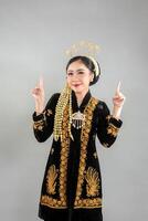 Young Asian woman wearing traditional Javanese attire with elegant golden embroidery, smiling confidently and pointing upward with both index fingers, showing joyful and inviting expression. photo