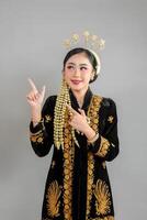 Beautiful young Asian woman wearing traditional Javanese kebaya and intricate golden accessories, smiling confidently while pointing upward with expressive, engaging facial expression. photo