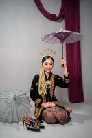 Asian woman in traditional Javanese costume smiles softly while holding a decorative umbrella. Her gentle expression highlights elegance, cultural pride, and classic feminine charm. photo