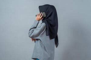 Side view of a young Asian Muslim woman wearing a black hijab and grey shirt, standing against a plain background while making a phone call using a smartphone, appearing calm and focused. photo
