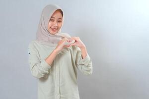 Smiling young Muslim woman in hijab making a heart shape with her hands while looking at the camera. Her cheerful facial expression radiates love, happiness, and kindness. photo