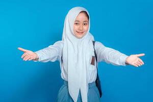 Cheerful Muslim schoolgirl in uniform and hijab smiling with open arms, welcoming warmly. Her happy facial expression conveys friendliness. Isolated on blue background. photo