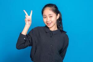 Smiling young Asian woman showing a peace sign with her fingers, radiating happiness and confidence. Her cheerful facial expression conveys friendliness and positivity on a blue background. photo