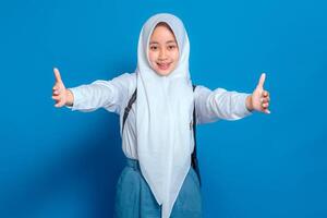 Cheerful Muslim schoolgirl in hijab smiling with open arms, expressing warmth and excitement. Her happy facial expression welcomes new opportunities. Isolated on blue. photo