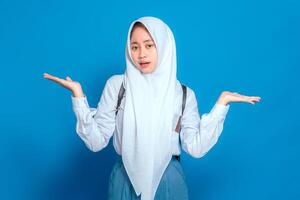 Confused Asian Muslim teenage girl in school uniform with backpack, shrugging with uncertain expression, isolated on blue background, back to school concept. photo