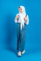 Confident Asian teenage girl in school uniform and hijab smiling gently while holding backpack straps, showing positive and proud facial expression, standing on blue background. photo