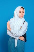 Confident Muslim schoolgirl in uniform and hijab showing stop hand gesture with calm, assertive facial expression. Back to school concept on isolated blue background. photo