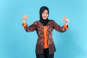 A cheerful young Asian woman in a hijab and batik outfit smiles while confidently pointing both index fingers downward, showcasing a playful and friendly facial expression in studio. photo