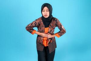 A young Asian woman in a batik outfit and hijab appears to be in discomfort or pain, holding her stomach with both hands while showing a concerned facial expression. photo