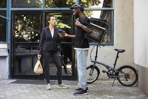Black delivery person completes a food transaction as customer pays with smartphone. The quick service interaction takes place during a city service drop off at her doorstep. photo