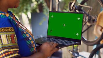 Detailed view of african american lady grasps minicomputer with greenscreen display as man in background removes broken wheel. Black woman holds laptop with blank mockup screen for bike maintenance. photo