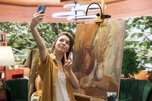 Confident painter self advertising her new masterpiece on online channel, filming a vlog on smartphone to promote her art skills for better reach and engagement. Influencer makes content creation. photo