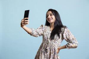 Joyful indian girl smiling at pictures and posting them online, feeling beautiful and confident in the studio. Excited upbeat woman taking photos using phone selfie camera, blue background.