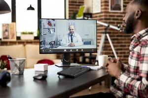 Black male employee seated at desk working remotely, describes symptoms during virtual consultation with caucasian doctor. Professionals discuss telemedicine project from home office via call. photo