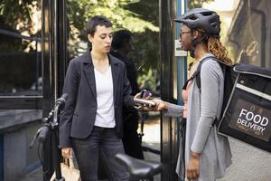 Black female worker accepts electronic payment from customer with a card on pos terminal. Courier ensuring transaction finalizes the purchase of fast food order from restaurant delivery. photo