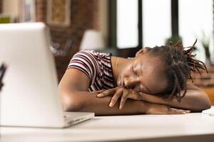 Black female entrepreneur lies on desk in home office, taking quick nap after demanding morning of work. Tired remote worker rests near laptop, overwhelmed by exhaustion from completing tight deadlines photo