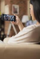 Back view of female employee on call with multiracial colleagues during online team briefing. Businesswoman seated on couch, attends virtual group meeting on mobile phone from home. photo