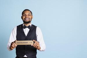 Doorman bellhop showing the restaurant direction with a hotel sign, holding an indicator to point guests to the amenities. Professional hotel concierge in suit and tie using a wall mark. photo