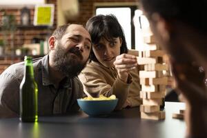 Young caucasian woman intensely focused on pulling wooden block from a jenga tower during tabletop game. Seated with snacks and drinks as her male friend watches the tense moment unfold. photo