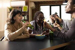Diverse friends talking, laughing and sharing snacks and beer during casual hangout in brick wall room. Multiethnic group seated around table, enjoying weekend downtime indoors with drinks and chips. photo