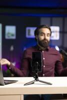 Joyful content creator sitting at table talking in front on smartphone on tripod in dimly lit home studio interior, chatting with fans. Man using mobile phone to record vlog in apartment photo