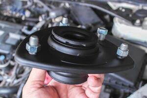 Top Strut Mount, A close-up of hand holds a black automotive strut mount with visible bolts over a car engine. photo