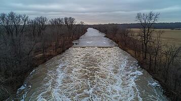 Aerial view of a river with a turbulent waterfall surrounded by bare trees under an overcast sky. photo