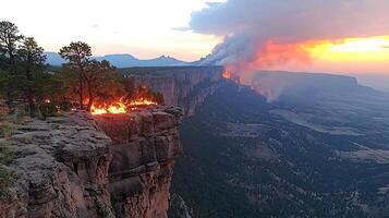 Wildfire rages across a dramatic cliff face at sunset. A massive plume of smoke billows into the sky, illuminated by the intense orange glow of the flames. photo
