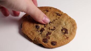 A hand breaks and picks up a chocolate chip cookie on a white surface under soft lighting video