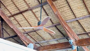A wooden ceiling fan spins under a straw roof in a tropical setting, with natural lighting highlighting the rustic architecture video