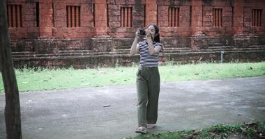 A young woman in a striped shirt and green pants walks along a path, taking photos with her camera. video