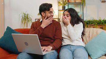 Cheerful young diverse couple in casual clothes using laptop and surprised with email winning lottery on sofa in living room at home. Happy Indian family giving high-five clenching fists in apartment. video