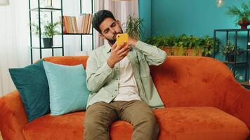 Sad displeased Arabian man use smartphone typing browsing, loses becoming surprised sudden lottery game results, bad news, fortune loss, fail, deadline, virus. Young Indian guy at home room on couch video
