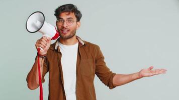 Indian young man talking with megaphone, proclaiming news, loudly announcing advertisement discounts sale, using loudspeaker to shout speech. Happy Arabian guy isolated on studio gray background video