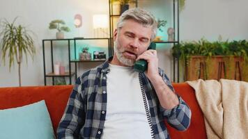 Sad senior man with gray hair talking on vintage retro wired telephone at home apartment. Disinterested Caucasian guy having annoyed boring conversation with hotline helpline service sitting on sofa. video