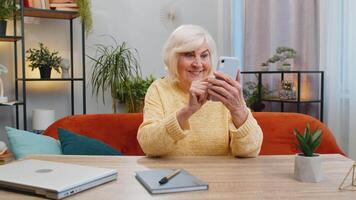 Happy senior woman use mobile smartphone typing browsing celebrating success victory, winning lottery jackpot achievement play game good positive news at home. Elderly mature grandmother sits on couch video