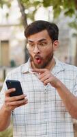 Oh my God, Wow. Excited happy joyful winner young man using smartphone pointing at screen browsing celebrating win good message news. Indian guy traveler with mobile phone in urban street outdoors. video