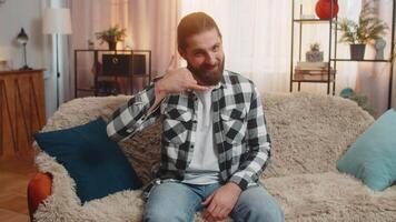 Young man sitting at home sofa with hand shaped like a phone, smiling in open anticipation for future call gesture asking for conversation. Caucasian guy waiting for contact from someone important. video
