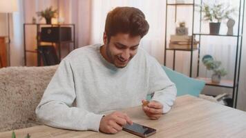 Young Indian man with bitcoin checks cryptocurrency exchange charts on smartphone app calculates approximate profits and shows BTC coin to camera celebrating win. Hispanic guy looks satisfied at home video
