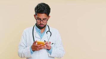Indian doctor cardiologist man using smartphone showing pointing empty place, advertising application area, positive feedback, copy-space. Arabian apothecary pharmacy guy isolated on beige background video