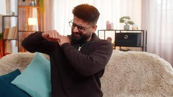 Indian man at home holds shoulder with tension, showing pain on his face while sitting on sofa. Arabian guy reacts quietly to soreness, trying to ease discomfort caused by muscle or joint strain video