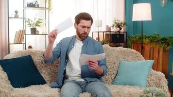 Young angry man at home desperately throws a pile of unpaid bills on the sofa, feeling overwhelmed. Caucasian guy clutches his head in frustration, struggling with financial debt and rising expenses. video