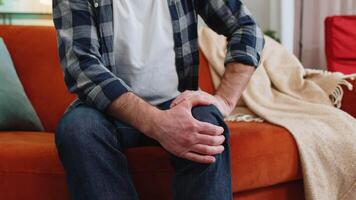 Unrecognizable middle-aged man suffer from painful discomfort severe knee joint ache leg muscles could not walk. Sick guy massaging knees illness cramps rheumatism at home on sofa. Health care problem video