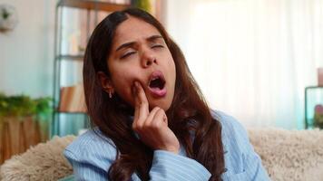 Indian young woman presses cheek in pain from sudden toothache while sitting on home sofa expression strained and tense. Hispanic girl shows clear dental discomfort and distress from oral health issue video