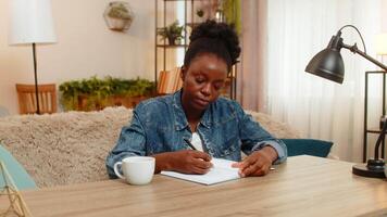 African American young woman sitting at table on home sofa thoughtfully sketches a new logo in notebook, deeply focused on creative design work. Black girl expresses artistic ideas with inspiration. video