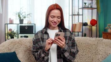Young happy woman at home scrolling news feed on smartphone with pleasant smile occasionally reacting to updates. Caucasian redhead girl on sofa enjoying social media content, relaxed cheerful moment video