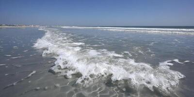 Ocean Wave Crashing on Sandy Beach Shoreline Buildings in Background Sunny Day Seascape photo