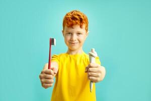 Red haired little boy holding brush tooth and tube of dental paste, preparing to clean teeth, wearing in yellow t shirt standing on blue isolated background. Sharpness on the hands, blurred face. photo