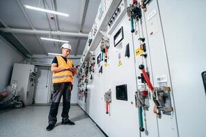 Adult electrician builder engineer testing and screwing equipment in fuse box and repairing modern electricity power station using data from the tablet. photo